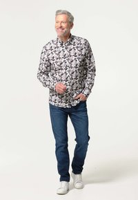 Lichtgekleurde button-up shirt met een bloemenpatroon, blauwe jeans en witte sneakers. Het model heeft een baard en kort haar.