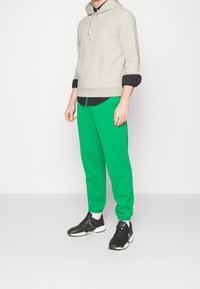 Szara bluza z kapturem z przodu z kieszenią i sznurkiem, w połączeniu z jasnymi zielonymi spodniami oraz czarnymi butami sportowymi z białymi akcentami.