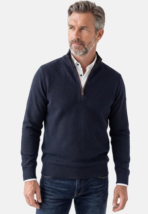 Mann mit grauem Haar und Bart, der einen marineblauen Pullover mit Viertelreißverschluss über einem weißen Hemd und blauer Jeans trägt und vor einem einfarbigen Hintergrund steht.