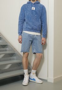 Sudadera de algodón azul deslavado con bolsillo frontal y logo; combinada con pantalones cortos de mezclilla claros con bordes deshilachados; zapatillas altas blancas y azules.