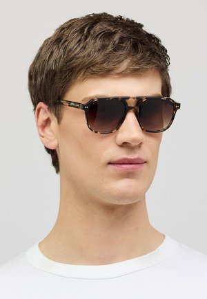 Jonge man met vierkante zonnebril met schildpadmotief en bruin verloopglas, gekleed in een wit shirt met ronde hals, tegen een neutrale achtergrond.