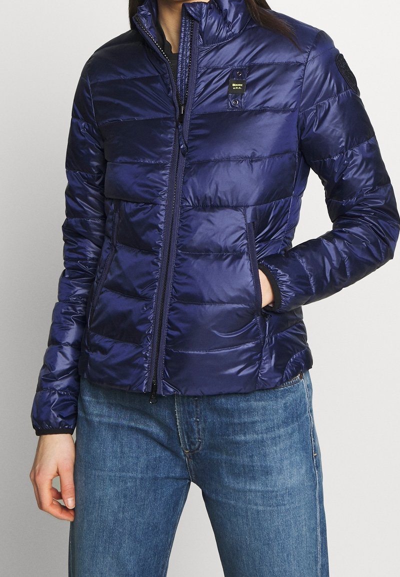Doudoune bleu marine avec fermeture éclair à l'avant, col montant et panneaux matelassés. Elle dispose de deux poches latérales et d'un patch logo sur la poitrine.
