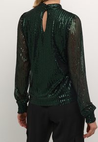 Grön paljettblus med hög halsringning, långa genomskinliga ärmar och detalj med nyckelhål baktill, med en figurnära fåll och strukturerat tyg.
