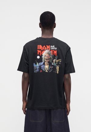 Homme portant un t-shirt noir Iron Maiden avec un graphisme d’un monstre grognant et le texte « Iron Maiden Est. 1975 », vu de dos.