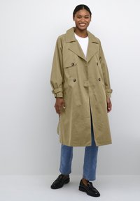 Beige Trenchcoat mit weitem Kragen, doppelt geknöpftem Design und zwei Fronttaschen. Mit umgeschlagenen Ärmeln gestylt und kombiniert mit blauen Jeans und Slippern.
