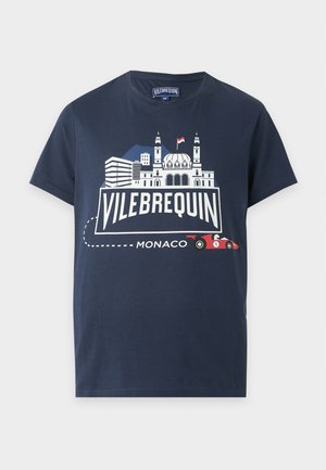 Homme portant un t-shirt graphique Vilebrequin Monaco bleu marine, un short beige et des sandales noires, debout les mains dans les poches sur fond blanc.