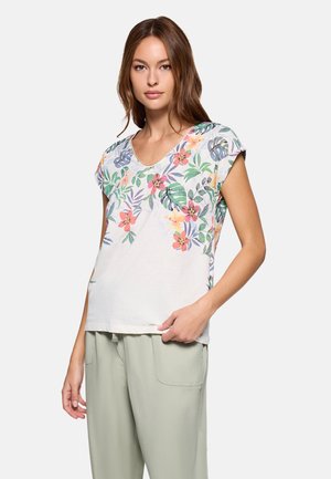 Mujer con una blusa blanca de manga corta con estampado floral tropical colorido y pantalones sueltos de color verde claro, de pie frente a un fondo liso.