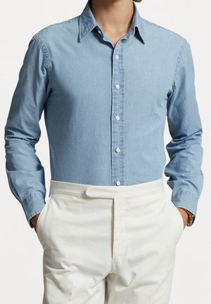Person trägt ein hellblaues Hemd mit Knöpfen, das in eine weiße, hoch taillierte Hose gesteckt ist, mit den Händen in den Taschen und einer braunen Uhr am linken Handgelenk.