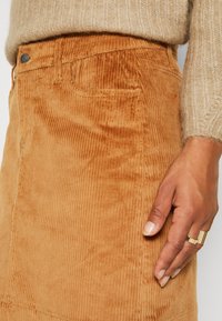 Gros plan d'une main avec des bagues en or reposant sur une jupe en velours côtelé beige, un pull en maille beige visible à la taille.