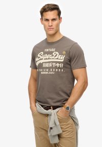 Camiseta gris de algodón con el logo "SuperDry" en blanco, cuello redondo, mangas cortas. Combinada con pantalones beige claros y un suéter gris atado a la cintura.