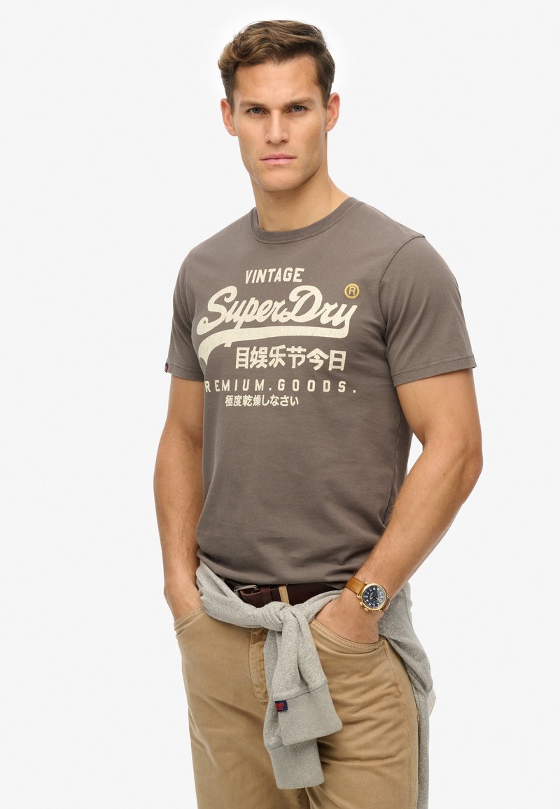 Camiseta gris de algodón con el logo "SuperDry" en blanco, cuello redondo, mangas cortas. Combinada con pantalones beige claros y un suéter gris atado a la cintura.