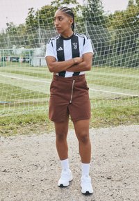 Schwarz-weiße gestreifte Adidas-Jersey kombiniert mit einem braunen Rock mit Reißverschluss. Tragen mit weißen Turnschuhen und Knöchelsocken. Grasfeld im Hintergrund.