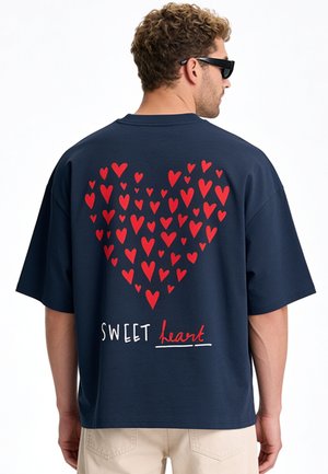 Mann trägt dunkelblaues T-Shirt mit großem roten Herz aus kleineren Herzen und den Worten "SWEET heart" auf dem Rücken.