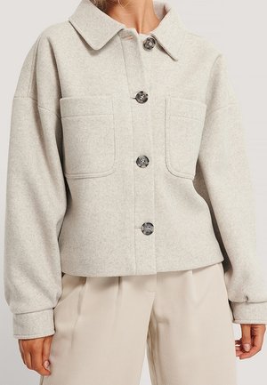 Lichtbeige wollen jasje met knopen, twee grote voorzakken, een brede kraag en lange mouwen, gedragen met bijpassende beige broek.