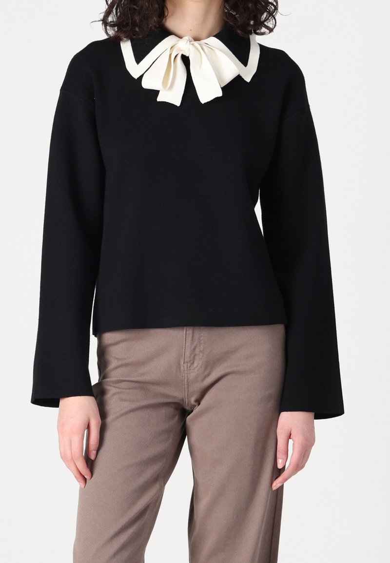 Femme portant un haut noir à manches longues avec un col à nœud papillon blanc et un pantalon taupe, debout devant un fond clair uni.