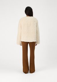 Krämfärgad, fluffig oversized-jacka i mysig textur, matchad med bruna ribbade flarebyxor, sedd från baksidan. Minimalistisk design utan synlig hårdvara.