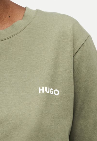 Gros plan sur une chemise vert olive montrant le logo blanc "HUGO" sur la partie gauche de la poitrine ainsi qu'une partie de l'encolure et de la couture de l'épaule.