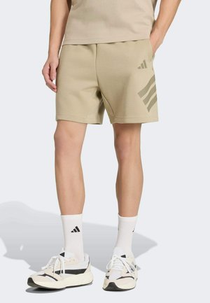 Beige Sportshorts mit weichem Stoff, lockerem Sitz und einem Drei-Streifen-Logo auf der linken Seite. Kombiniert mit weißen Socken und Turnschuhen.