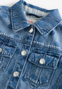 Veste en denim de couleur bleu clair, avec de grands pois bleu foncé, deux poches avant, des boutons pression argentés et des coutures décoratives.