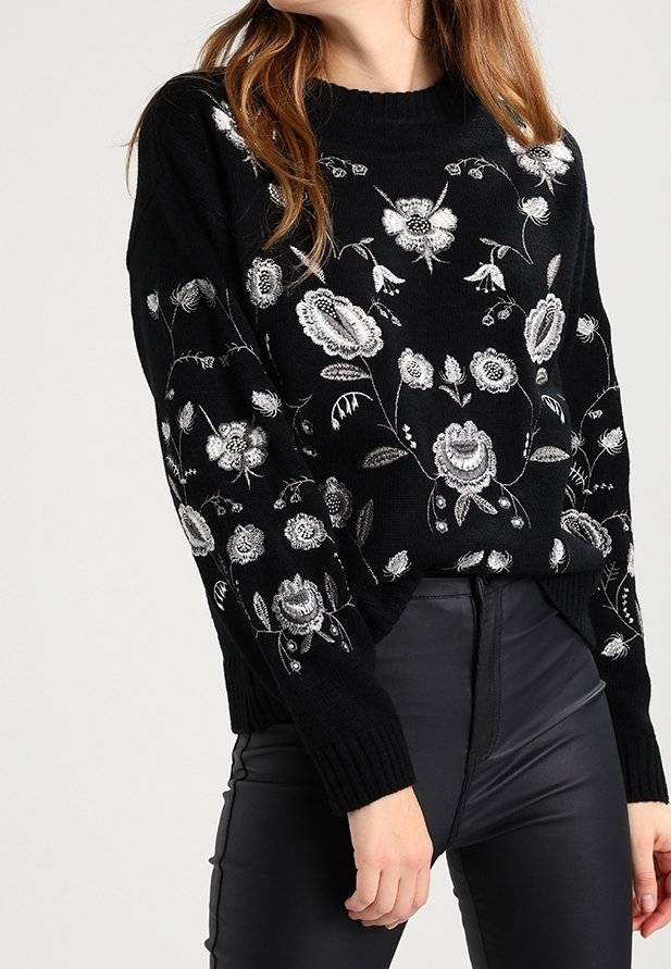 Pull en maille noir avec broderie florale blanche porté avec un pantalon noir en simili cuir taille haute, visible des épaules aux hanches.