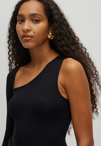 Zwarte body met een asymmetrische halslijn, gemaakt van gladde stof; model draagt grote, gestructureerde gouden oorbellen. Kroeshaar omlijst het gezicht.