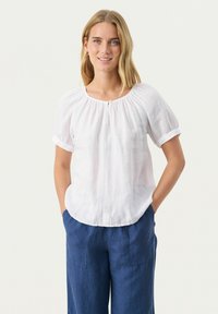 Weiße Leinenbluse mit kurzen umgeschlagenen Ärmeln, runder Halslinie und einem dekorativen Knopf vorne. Kombiniert mit blauen Leinenhosen.
