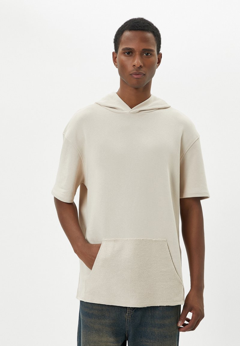 Beige huppari, jossa on lyhyet hihat, etutasku ja teksturoitu helma, valmistettu pehmeästä puuvillakankaasta.