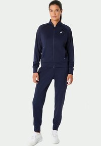 Navy tracksuit made of soft, stretchy fabric. Jacket features a full zip, high collar, and side pockets. Pants have a tapered fit and elastic cuffs.