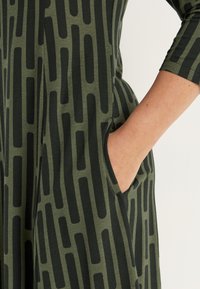 Vihreä mekko pehmeästä materiaalista, jossa on musta abstrakti kuvio, sivutasku ja kolmanneksen mittaiset hihat.