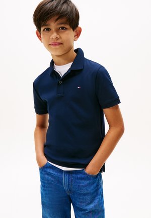 Ragazzo con capelli castani corti che indossa una polo blu navy e jeans blu, in piedi con le mani nelle tasche contro uno sfondo bianco.
