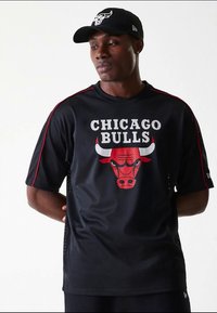 Melnā sieta Čikāgas "Bulls" krekls ar sarkano bulli logo un balto uzrakstu. Akcenti ietver sarkanas sānu svītras un brīvu piegriezumu.