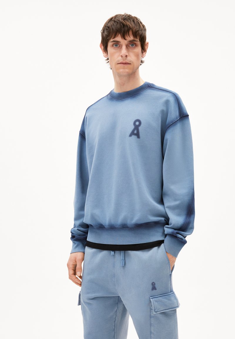 Man in een blauwe sweatshirt en bijpassende cargobroek met een lusvormig sleutelgatlogo op de borst en dij, staand tegen een witte achtergrond.