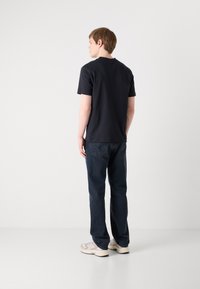 Krátký rukáv modrého trička, volný střih, kombinovaný s tmavě modrými džínami. Model má na sobě světlé tenisky, ukazuje ležérní outfit.