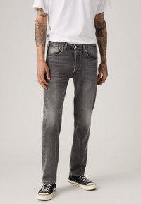 Man in vervaagde zwarte jeans, zwart-witte sneakers, een wit T-shirt, met zichtbare getatoeëerde armen, staande tegen een effen achtergrond.