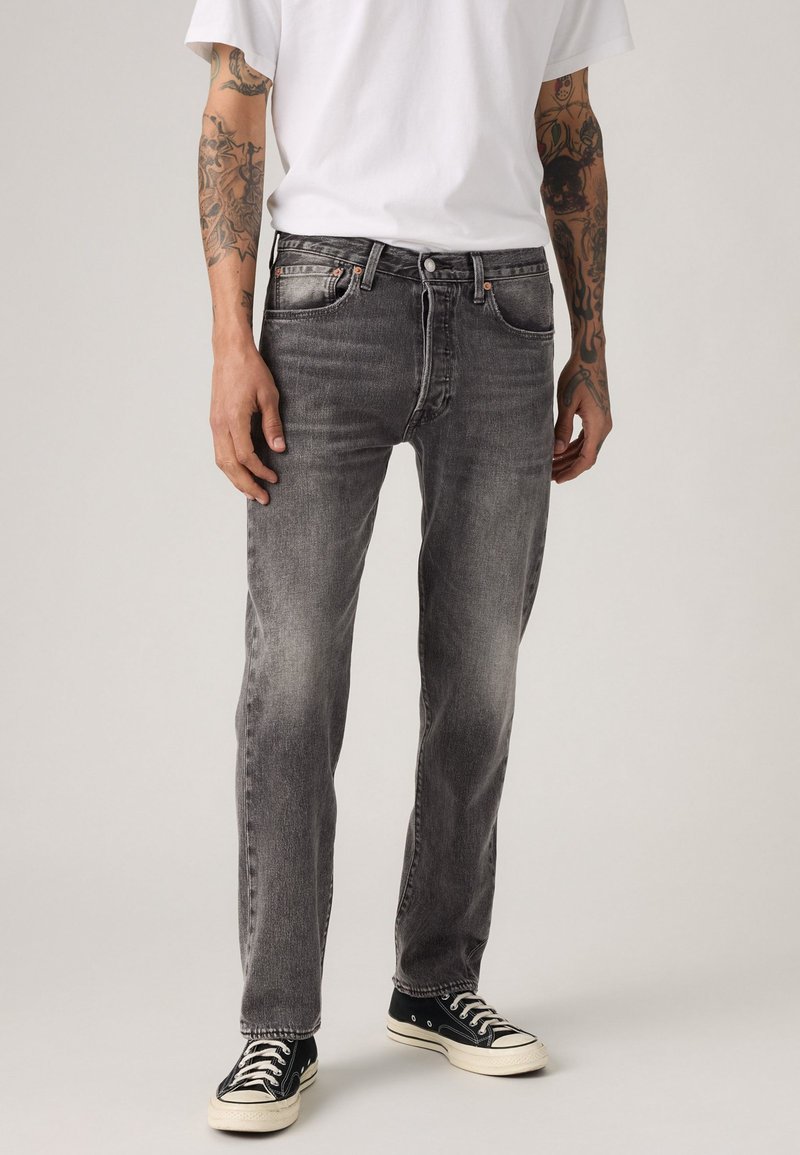 Man in vervaagde zwarte jeans, zwart-witte sneakers, een wit T-shirt, met zichtbare getatoeëerde armen, staande tegen een effen achtergrond.