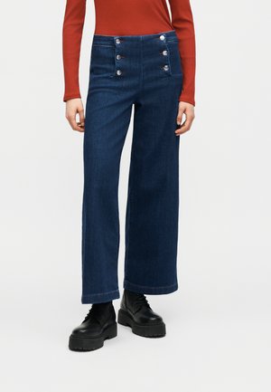 Σκούρο μπλε τζιν παντελόνι με ψηλή μέση και φαρδιά μπάρα, με έξι ασημένια κουμπιά μπροστά, φορεμένο με μαύρα χοντρά μποτάκια και μακρυμάνικη μπλούζα σε χάλκινο χρώμα.