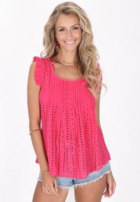 Rosa ärmelloses Oberteil mit Rüschenschultern, ausgestattet mit einem strukturierten Design und einem Lochmuster. Kombiniert mit Jeansshorts, was den lässigen Stil betont.