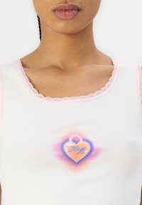 Hvit tanktopp med rosa blondedetaljer, med et fargerikt hjertegrafikk med "Hugo"-tekst i midten. Glatt stofftekstur.