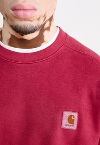 Felpa color bordeaux con collo a girocollo a costine. Presenta una toppa quadrata con logo in rosa e marrone, realizzata in tessuto morbido e testurizzato.