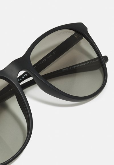 Occhiali da sole neri con finitura opaca, lenti grigie e montatura arrotondata. Presentano dettagli del marchio incisi sulle aste.