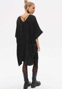 Schwarzes oversized Kleid mit tiefem V-Ausschnitt hinten, lockeren Ärmeln und seitlichen Schlitzen. Hergestellt aus weichem Stoff. Kombiniert mit schwarzen Knöchelstiefeln.