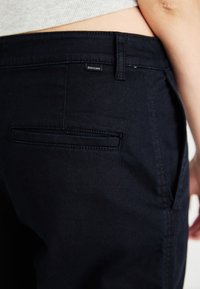 Dark blue pants made of cotton blend fabric. Features a back pocket and a small black logo tag. Smooth texture and tailored fit.