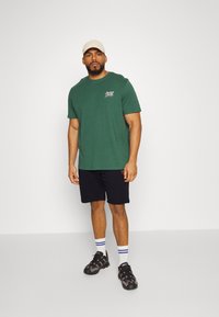 Green cotton t-shirt with short sleeves, featuring a small logo on the chest, paired with black shorts and black sports shoes with purple accents.
