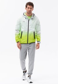 Giacca leggera con zip in gradiente da bianco a verde lime, caratterizzata da dettagli neri e due tasche con zip. Abbinata a pantaloni della tuta grigi e sneaker.