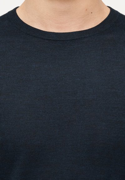 Κοντινό πλάνο στον λαιμό και το άνω μέρος του στήθους ενός ατόμου που φοράει πουλόβερ πλεκτό με στρογγυλή λαιμόκοψη σε σκούρο ναυτικό μπλε χρώμα.