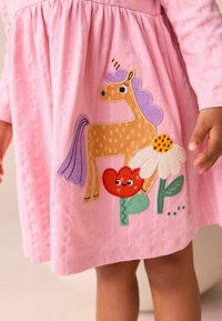 Różowa bawełniana sukienka z haftowanym jednorożcem o fioletowej grzywie, w otoczeniu kolorowych kwiatów, które dodają projektowi zabawnych akcentów.