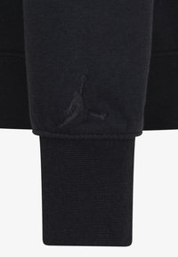 Polsino di una felpa nera realizzato in tessuto a maglia testurizzato. Logo ricamato di una figura che salta in nero abbinato vicino alla cucitura.