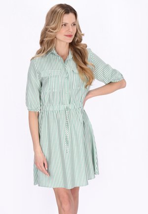 Kvinde med langt blondt hår iført en grøn og hvid vertikalt striped kjole med bælte og halværmer, stående med den ene hånd på hoften.