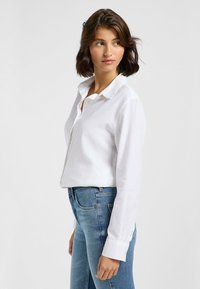 Weißes Leinen-Hemd mit Knopfleiste, lockerer Passform und spitzem Kragen, kombiniert mit mittelblauen Jeans. Verfügt über lange, hochgekrempelte Ärmel.