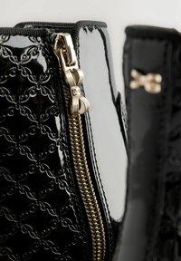 Schwarze Lackleder-Ankle Boots mit einem dekorativen geprägten Muster, ausgestattet mit einem goldenen Reißverschluss, der durch einen schleifenförmigen Zuggriff akzentuiert wird.